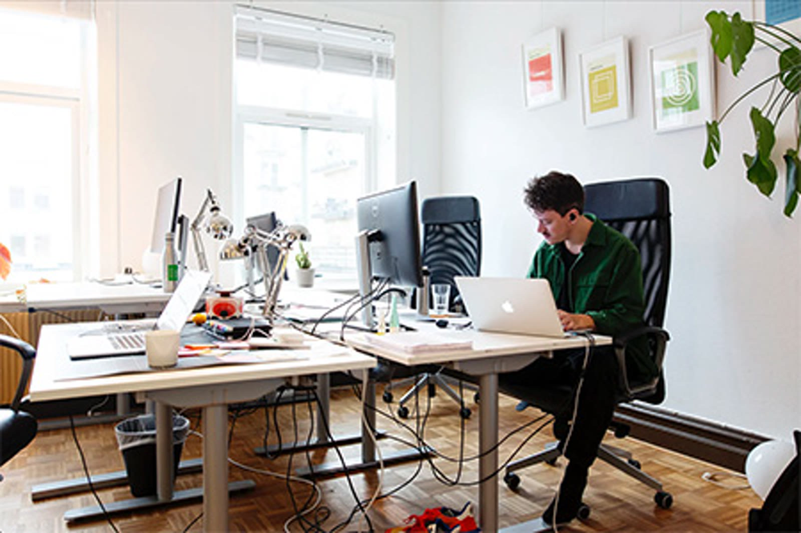 A founder working at a desk with a laptop, surrounded by colorful wall frames and natural Nordic light through large windows at The Ground