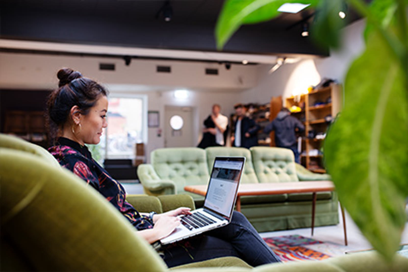 A founder working from a laptop on a velvet green sofa at The Ground, with a large monstera leaf in the foreground and other members in the background of the Living Room