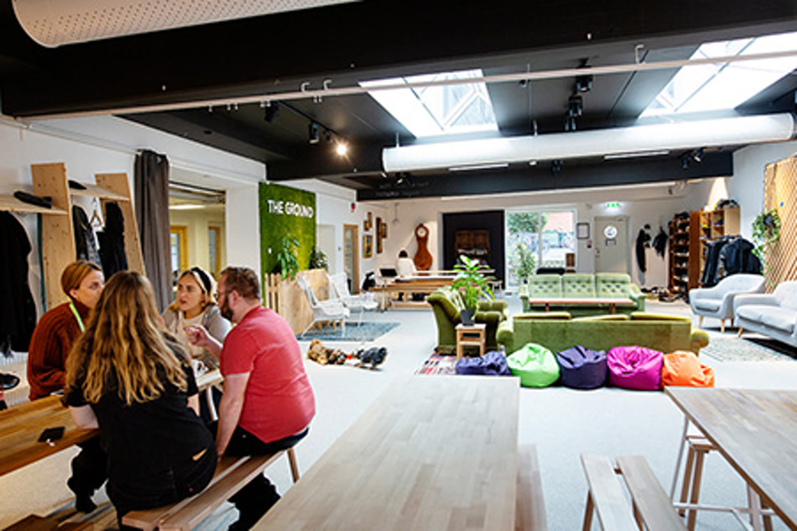 Wide view of The Ground Living Room — a long wooden table with members eating lunch together, pink beanbags on the floor, and the green THE GROUND wall logo in the background